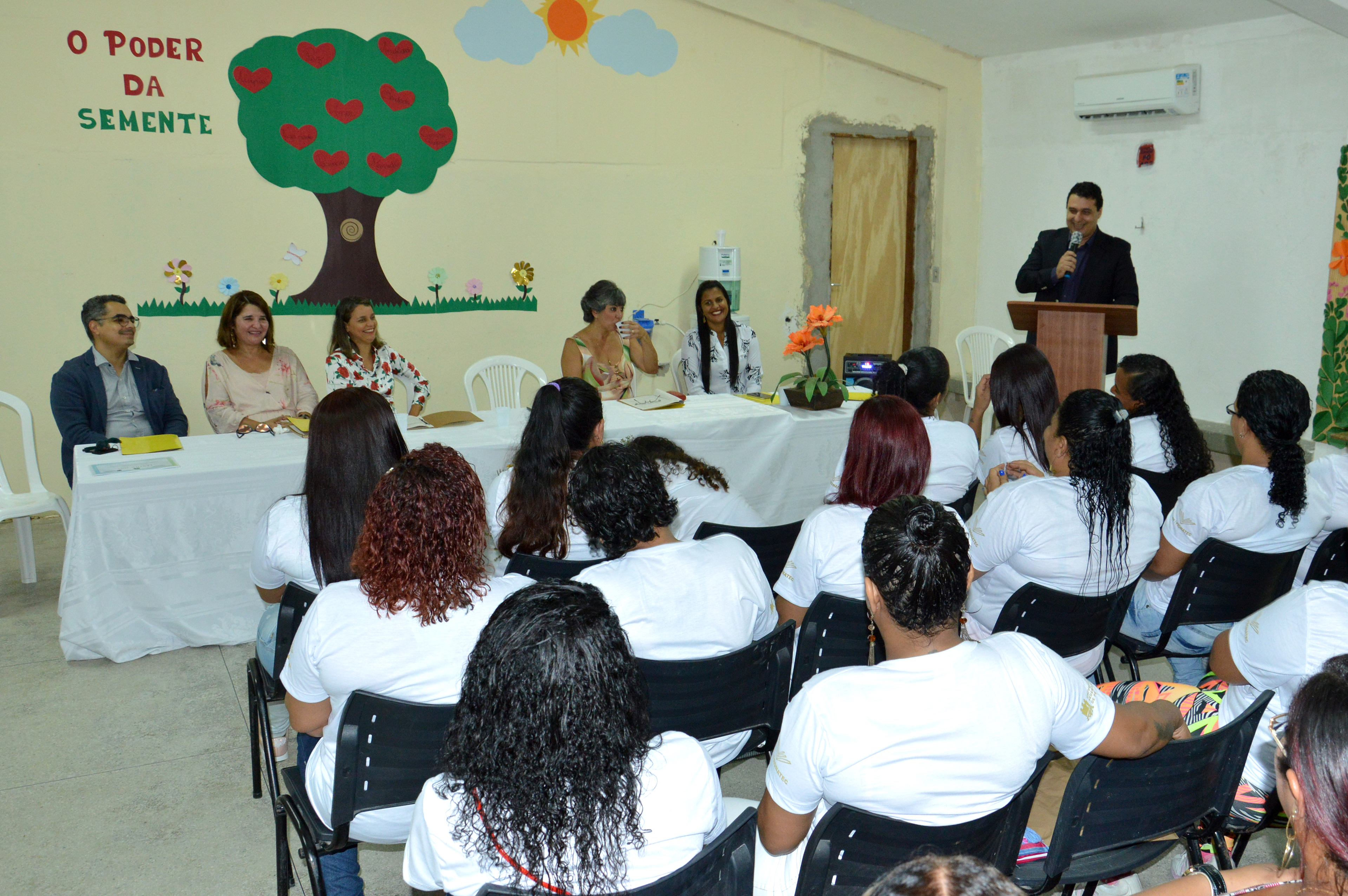 imagem de mulheres sentadas em cadeiras, ângulo por trás. à sua frente, professores e professoras do curso sentados numa mesa. enquanto um homem fala ao microfone num púlpito