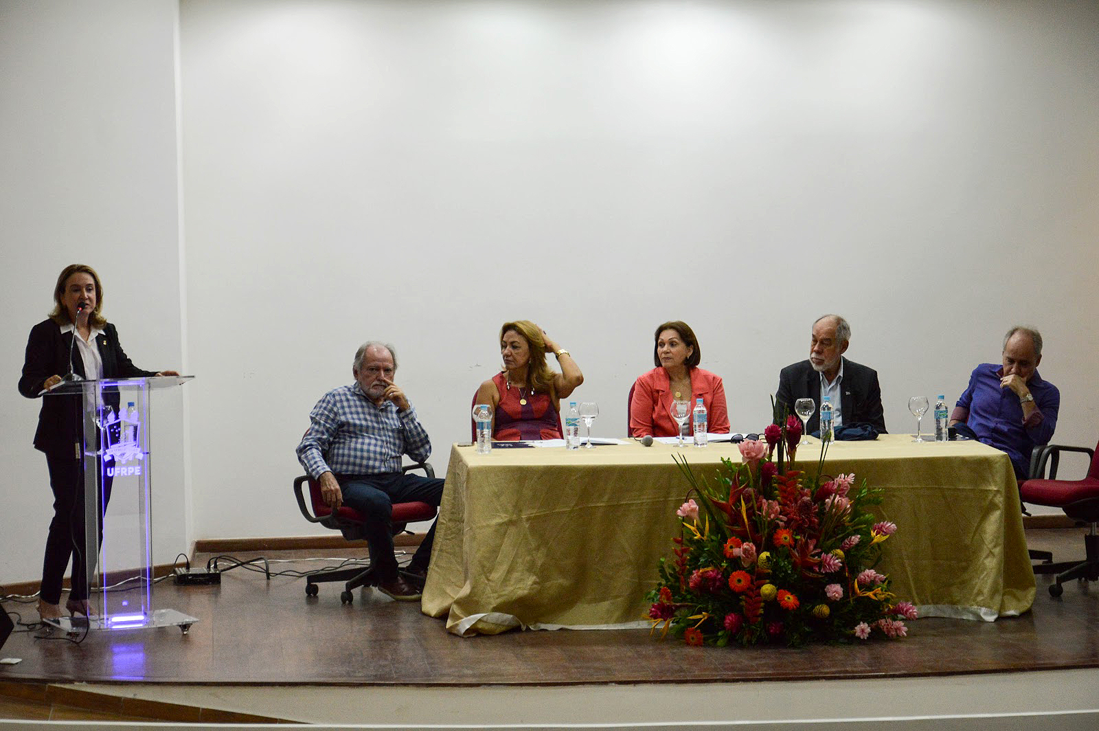 pesquisadores sentados à mesa de abertura do workshop, enquanto pró-reitora fala ao microfone