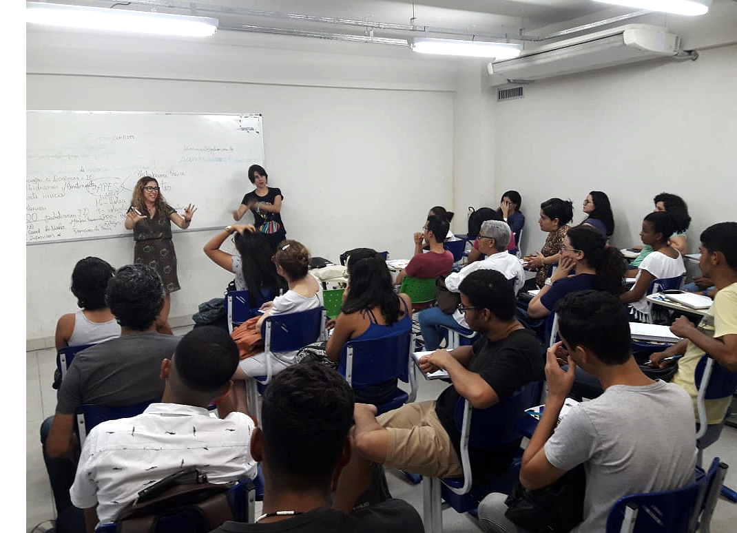 Estudantes do curso de Letras participam de aula, sentados em suas bancas. À frete, professora e interprete de libras ministram a aula